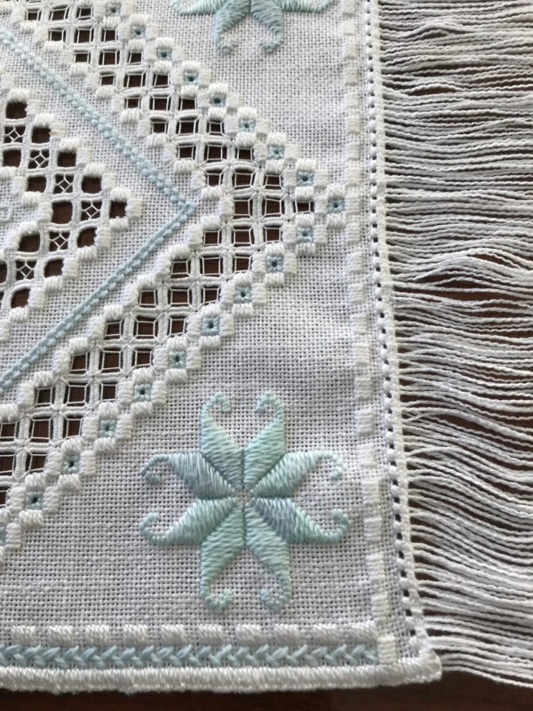 Photo of a small area of an embroidered placemat stitched in the Hardanger technique and showing the details of the stitches in the open areas and one of the repeating Hardanger flower motifs.