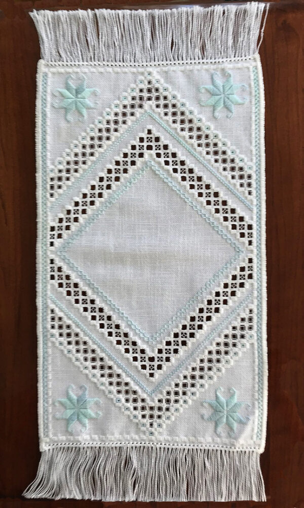 Photo of an embroidered placemat with fringe at each short edge, stitched in the Hardanger technique with open areas where threads have been cut after stitching and motifs that follow the grid made up by the fabric threads.