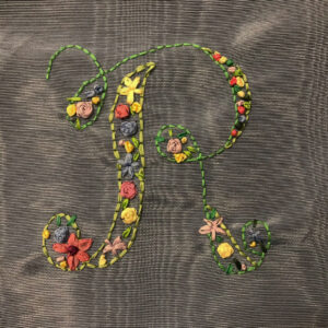 Photo of an script style uppercase "r" outlined ribbon embroidery and filled various ribbon flowers and leaves.