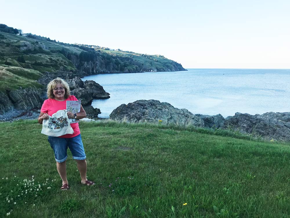 Photo of a woman on a grassy area with a rocky shoreline and water in the background. The owman is holding a magazine and some embroidery.