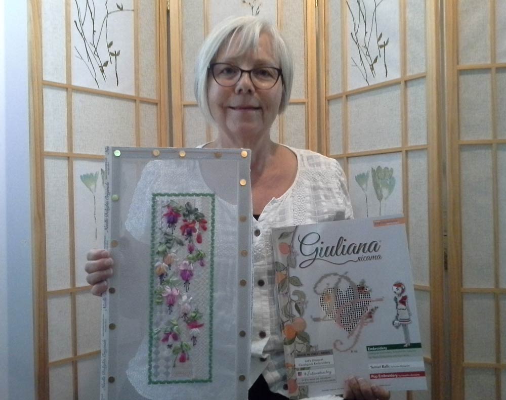 Photo of a woman holding a copy of the Giuliana Ricama magazine and a piece of canvas work enbroidery with colourful fuchsia flowers stitched with silk ribbon