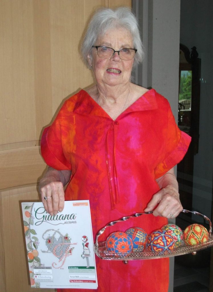 photo of a woman in a bright red dress holding a copy of Giuliana Ricama magazine and a basket with five temari - balls with surface patterns made with thread.