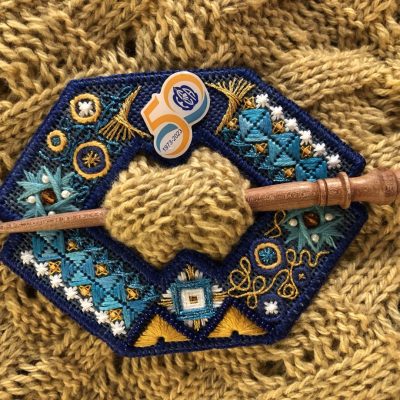 Photo of a shawl pin made from an embroidered geometric shape with a void in the centre and with a 50th-anniversary pin attached. It is held on a knitted fabric with a wooden skewer.