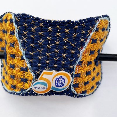 Photo of a hair holder made from an embroidered geometric shape with a 50th-anniversary pin attached. It has a wooden skewer that goes through an opening on each side to hold it in the hair.