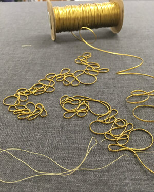photo of a spool of gold embroidery metal on a fabric as it is being formed into squiggly piles and attached to a piece of brown fabric