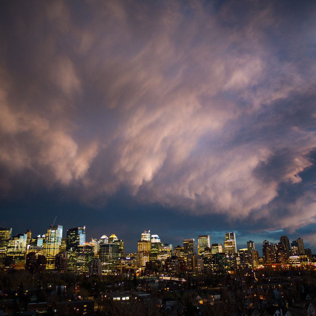 A photo of the Calgary skyline at dusk. The lights are turned on in the buildings. The sky is covered with clouds, but blue sky shows in the distance in a phenomenon known as a Chinook Arch.