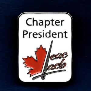 Photo of an enameled pin with a white background. The words Chapter President are at the top. There is a partial red maple leaf on the left. A gray sewing needle is dividing the maple leaf from the red letters "EAC" and "ACB" that are stacked on the right. A line representing a wide gray thread is under all of the elements. It looks like the thread is through the eye of the needle.