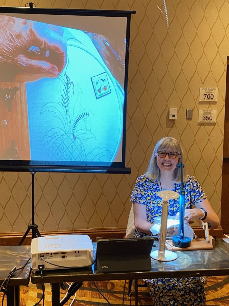 A photo of the instructor Kathy Andrews with a screen behind her sharing a close-up image of her stitching during a class at Seminar 2023.