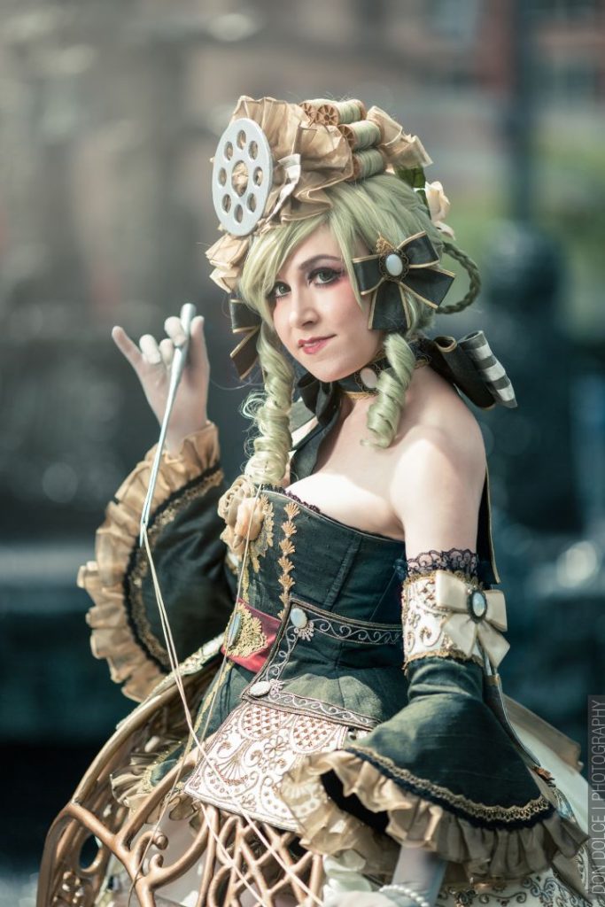 Cosplay woman character with pale green hair in an elaborate , partial up-do featuring spools in the curls, with an off-the-shoulder green, elaborately embroidered dress and a curlicued metal-looking hoop exposed at the front. Photo © Don Dolce Photography.