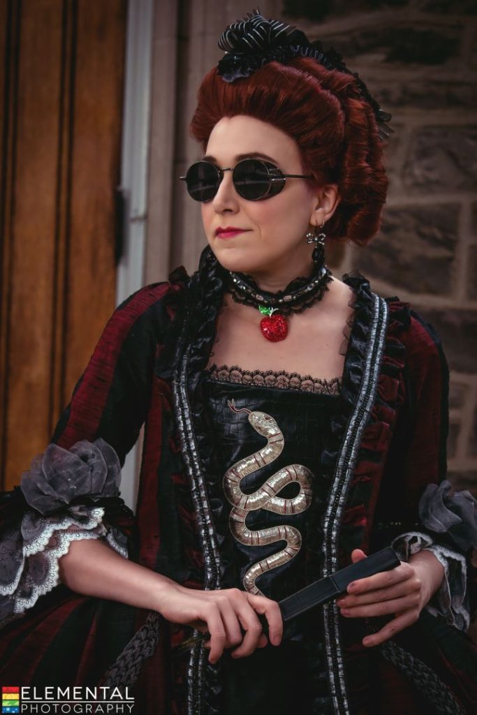 Cosplay character with dark auburn hair wearing dark, retro sunglasses, a sequined apple choker-style necklace, and a late 1800s style red and black striped dress with a stumpwork serpent on the bodice and elaborate trims.