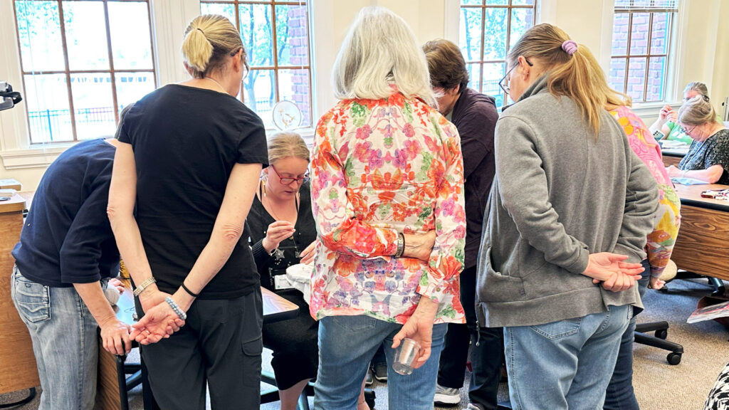 photo of a group of people gathered around an instructor who is demonstrating an embroidery technique