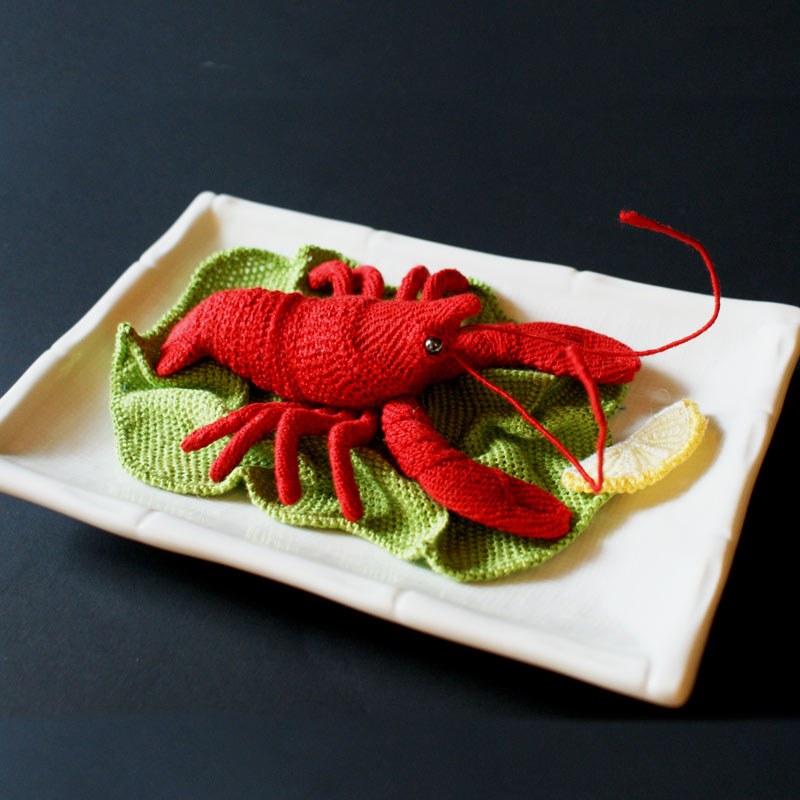 A photo of a plate with an embroidered, three-dimensional lobster on a bed of embroidered greens and with an embroidered lemon slice.