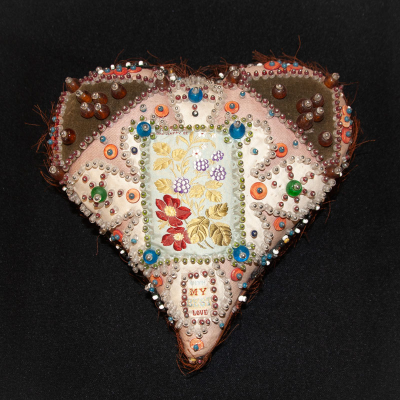 Photo of a heart-shaped pincushion with different fabrics making up the front and with the patches and seams embellished with embroidery and beads. The edges of the pincushion are frayed.