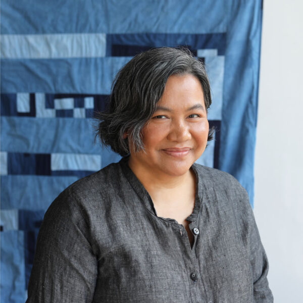 A photo of a woman's upper body in front of a backgraounf of blue fabric pieces sewn together.