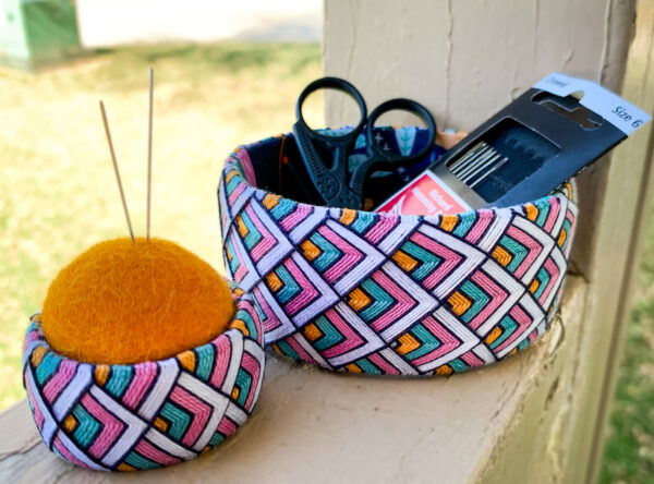 Photo of a open-topped round vessel with small sized scissors and packages of needles inside, and a smaller round vessel with a felt ball inserted and filling the opening with pins stuck into it.