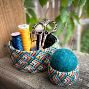 Photo of a open-topped round vessel with spools of thread and small sized scissors, and a smaller round vessel with a felt ball inserted and filling the opening with pins stuck into it.