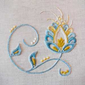 Photo of an embroidery of a swirled fantasy flower, stem and leaves in the Jacobean style stitched with blues and yellows.