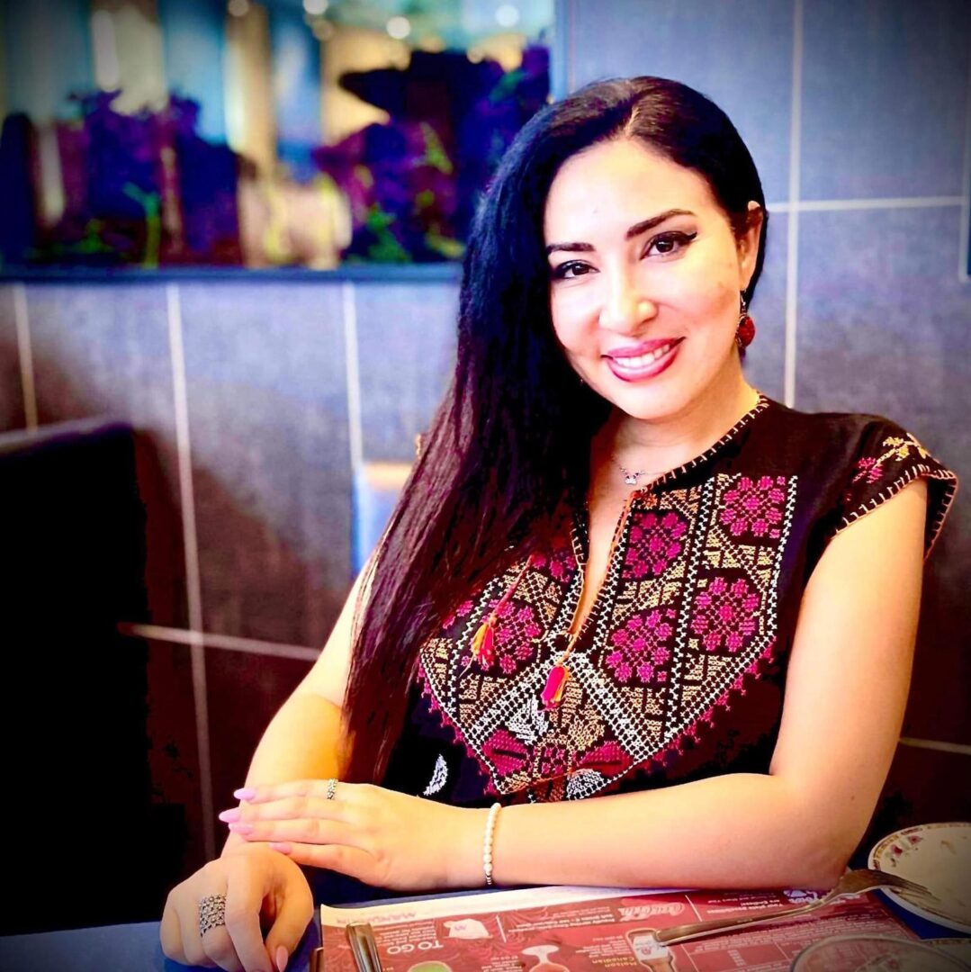 Photo of a sitting woman with long dark hair wearing a dark sleeveless tunic with colourful tatreez-style embroidery on the yoke and along the edges of the arm and neck openings.