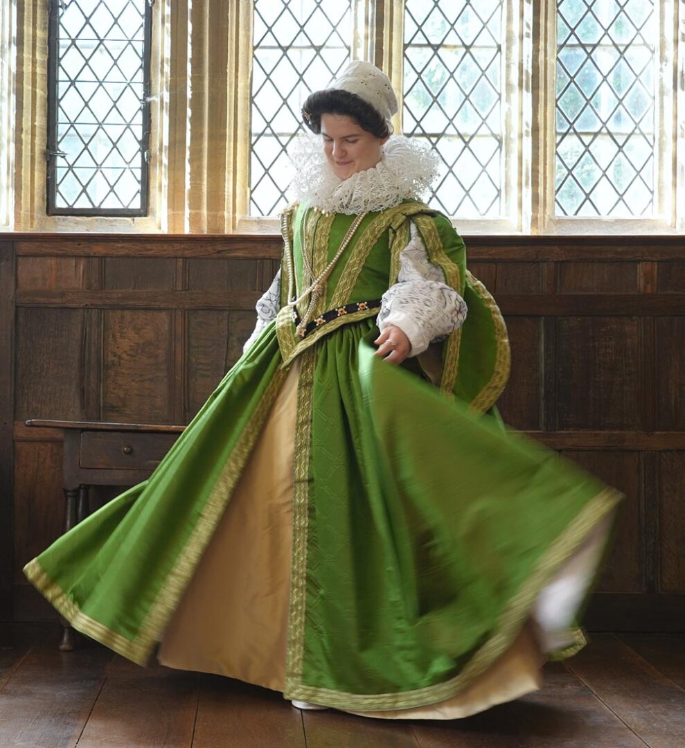 Photo of a woman in an historic-style full length dress in green with an embroidered bodice, a gold underskirt and gold trim. There are full puffy sleeves and a neck ruff.