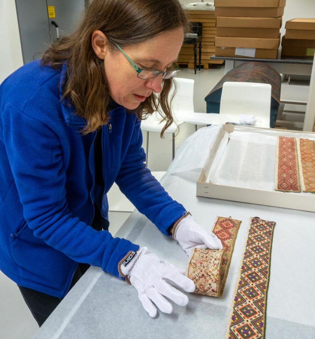 A photo of a woman wearing white gloves at a table, with a box of embroidered strips to her left, shows her folding back a strip of embroidery to look at the back.