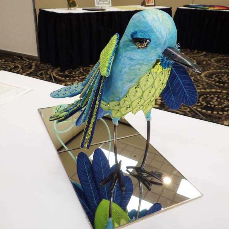 Photo of a three-dimensional bird made with embroidered pieces of fabric for the body, wings and tail, and metal for the beak, legs and feet. It has elaborately embroidered wings, tail and breast, and fancifully long eyelashes. The makers named it "Avis."