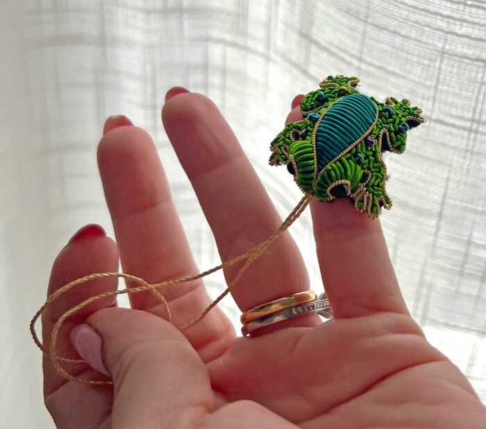 Photo of a tiny frog's back, legs, toes and head, embroidered with metal threads over a shaped form, sitting on a person's smallest finger. 