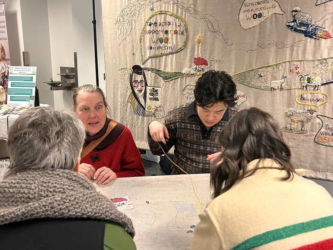 Four people sit around the table, 3 are currently talking while a gentleman continues to stitch. Behind them hangs part of the tapestry. On the tapestry it shows a woman holding a book and saying "support your wool community" and beside her are sheep and hills, and various other stitched items.