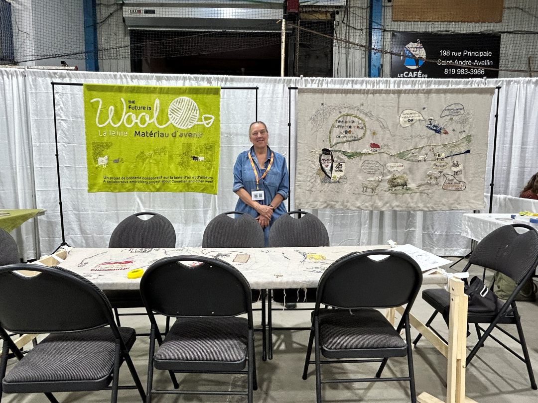 Speaker Kathleen Vaughan stands behind a stand on which part of her presentation canvas is stretched. Behind her is a sign that reads "The future is Wool" and on the other side is a canvas on which is stitched various quotes, sheep, a lady and other items.