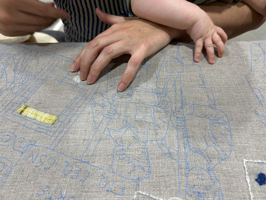 This photos shows two hands...mother and child most likely, resting on the canvas while the mother stitches in white yarn. The canvas has the outline of what is to be stitched in blue. The blue lines show an image of 2 people signs which we cannot read.